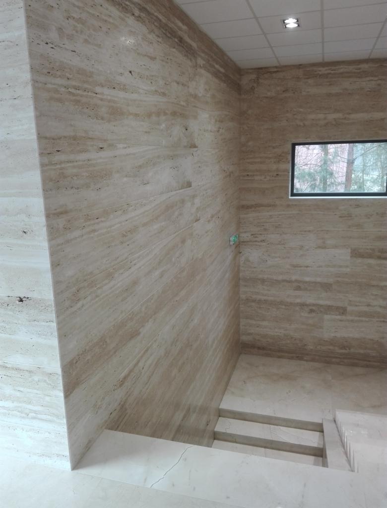 Wnętrze wykończone trawertynem: ściany i schody w odcieniach beżu i brązu, małe okno z widokiem na drzewa.