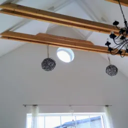 Wnętrze z wysokim, skośnym sufitem, drewnianymi belkami stropowymi, okrągłym oknem dachowym i ozdobnymi kulami zwisającymi na sznurkach, widoczny żyrandol z czarnego metalu.