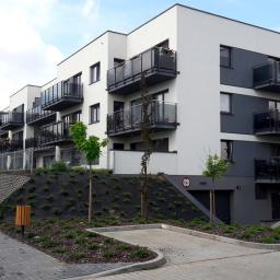 WYKOREM Sp. z o.o. - Nowoczesny budynek wielorodzinny z balkonami i elewacją w kolorach białym i szarym, widok z zewnątrz na uporządkowany teren z młodymi drzewkami i chodnikiem z kostki brukowej.