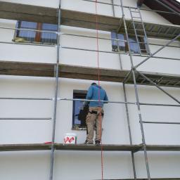 WYKOREM Sp. z o.o. - Pracownik na rusztowaniu maluje białą elewację domu jednorodzinnego, widoczny kubeł z farbą Kabe i zabezpieczone okna.