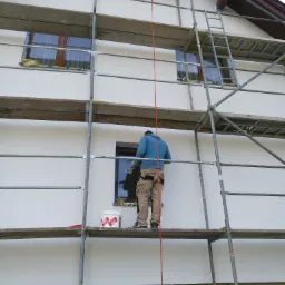 Pracownik na rusztowaniu maluje białą elewację domu jednorodzinnego, widoczny kubeł z farbą Kabe i zabezpieczone okna.