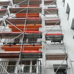 WYKOREM Sp. z o.o. - Wysokie rusztowanie metalowe na tle jasnej elewacji budynku wielorodzinnego, widoczne balkony z pomarańczowymi elementami wykończeniowymi.