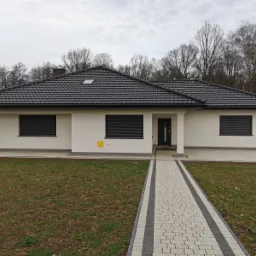 Nowoczesny dom jednorodzinny z ciemną dachówką i jasną elewacją, widoczne rolety zewnętrzne na oknach i ścieżka z kostki brukowej prowadząca do wejścia, w tle drzewa bez liści.