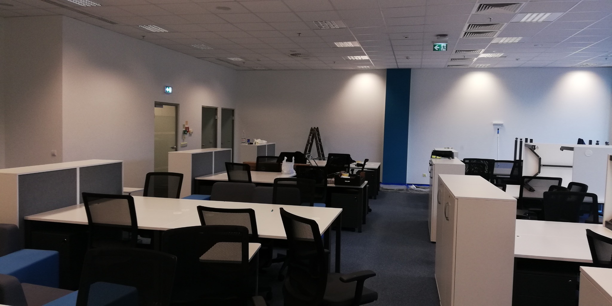 Przygotowane do użytku, odświeżone biuro w Ćwiklicach z białymi biurkami i czarnymi krzesłami, widoczne ślady malowania ścian i sufitu, pozostawione drabiny malarskie.