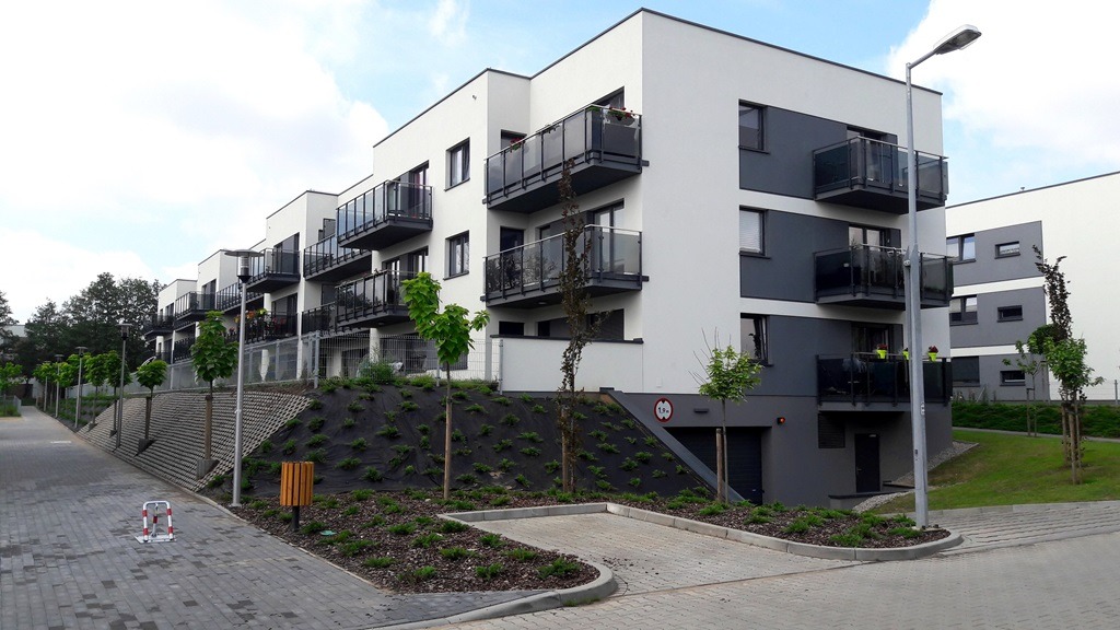 Nowoczesny budynek wielorodzinny z balkonami i elewacją w kolorach białym i szarym, widok z zewnątrz na uporządkowany teren z młodymi drzewkami i chodnikiem z kostki brukowej.