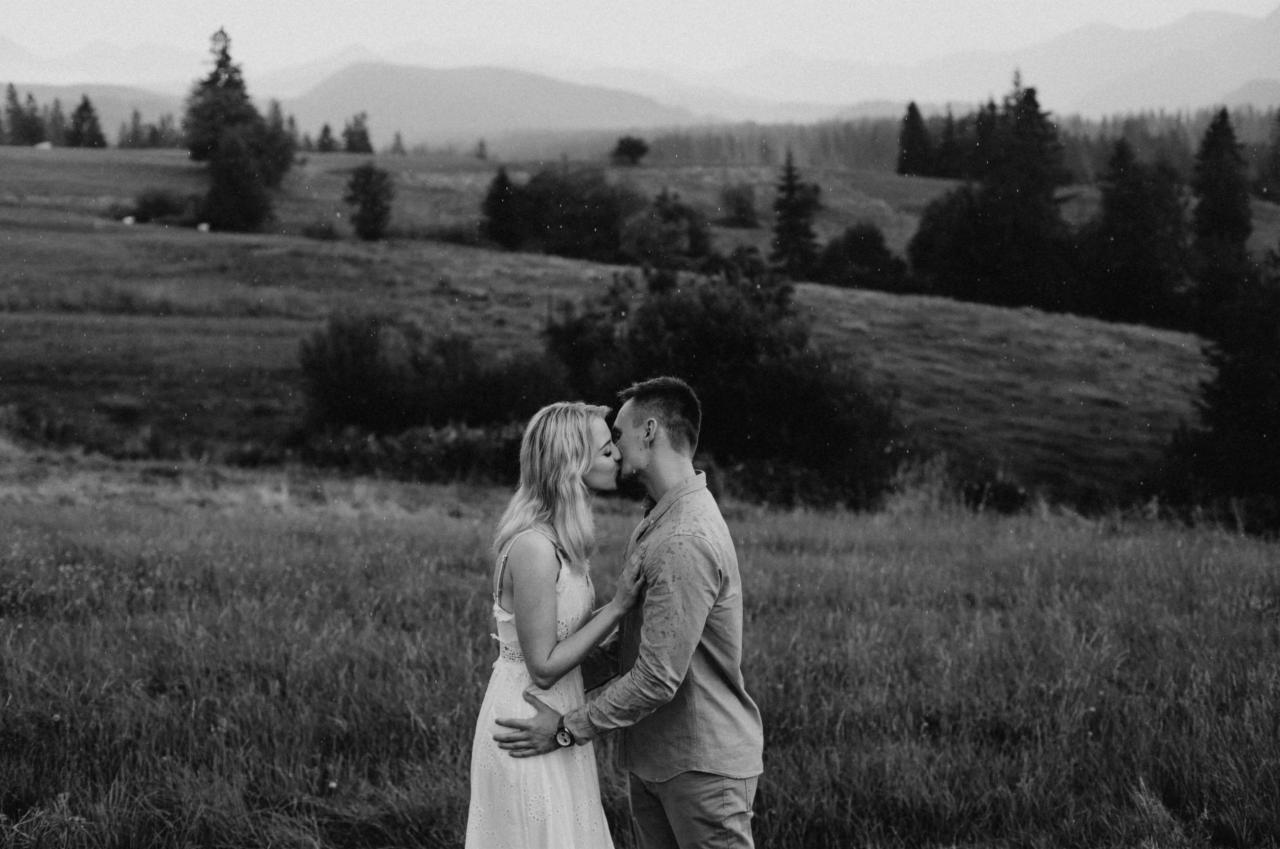 Czarno-białe zdjęcie całującej się pary na tle łąki i gór; mężczyzna w koszuli z długim rękawem obejmuje kobietę w białej sukience.