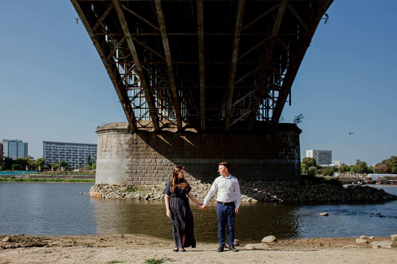 Para trzymająca się za ręce pod masywnym, żelbetonowym mostem nad rzeką, z budynkami w tle i piaskiem na brzegu.