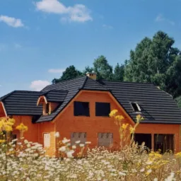 Surowy drewniany dom w stanie deweloperskim z ciemnym dachem z dach&oacute;wki metalowej, otoczony łąką kwitnących kwiat&oacute;w i drzew, na tle błękitnego nieba.