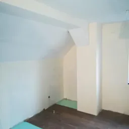 Pokój na poddaszu w trakcie układania paneli: widoczna zielona pianka podkładowa i ułożone panele podłogowe w odcieniach brązu, ściany pomalowane na jasny kremowy kolor.