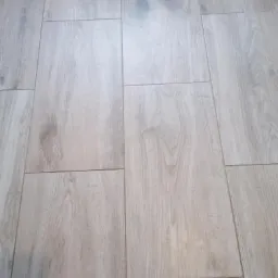 Ułożone podłużne płytki podłogowe imitujące jasne drewno, z ciemniejszymi akcentami w słojach, wypełnione fugą w kolorze zbliżonym do płytek.