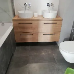 Nowoczesna łazienka z dwiema okrągłymi umywalkami nablatowymi na drewnianej szafce z czarnymi uchwytami, wisząca toaleta i szare płytki podłogowe.