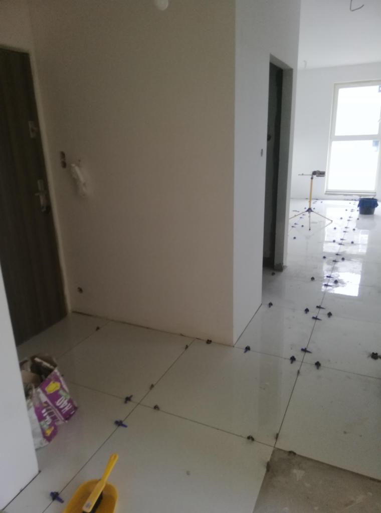 Układanie białych płytek podłogowych w jasnym wnętrzu, widoczne krzyżyki dystansowe, wiadro i torba z klejem, ślady po starej posadzce.