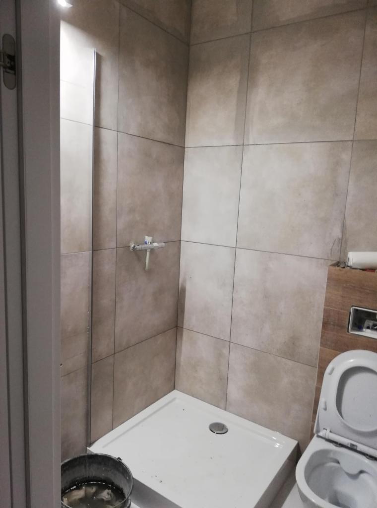 Nieduża kabina prysznicowa z brodzikiem i zamontowaną baterią, wykończona płytkami w odcieniach beżu, z widocznym wiadrem z wodą i otwartą toaletą.