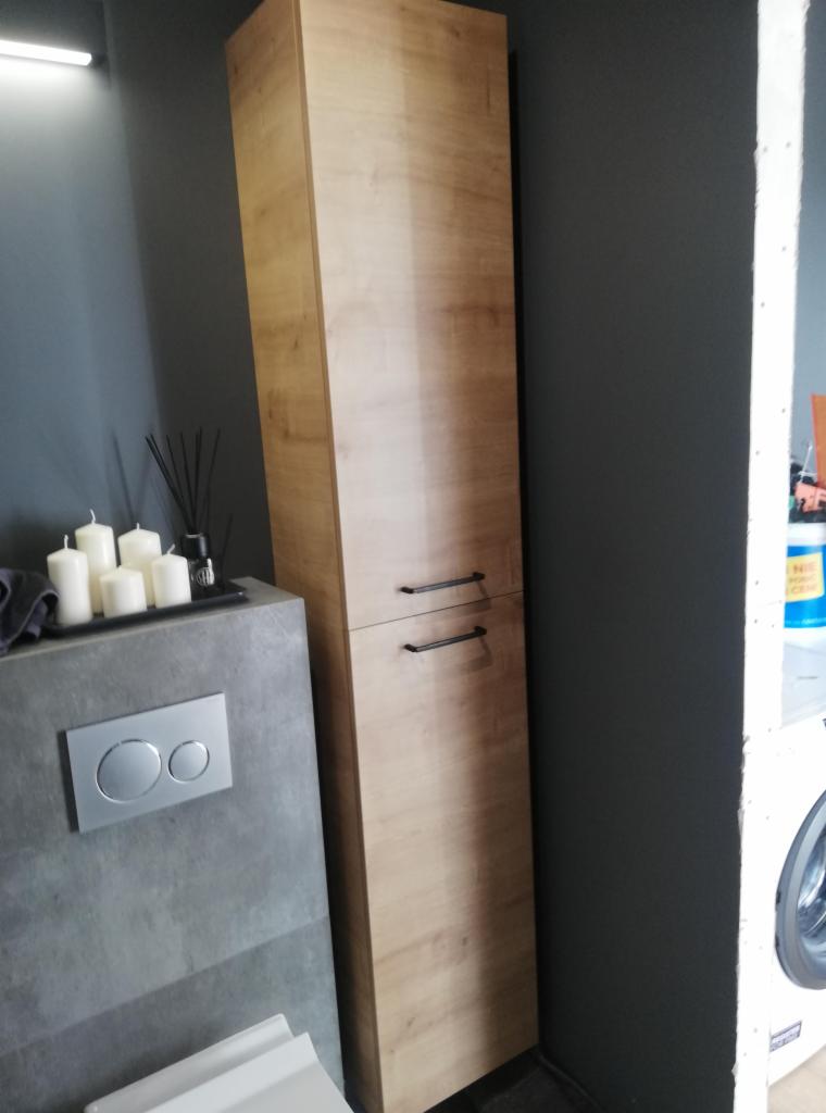 Wysoka, wąska szafka łazienkowa z drewnopodobnym fornirem i czarnymi uchwytami, obok szarej ściany, umywalki z szarą obudową i białej miski WC, w tle pralka.