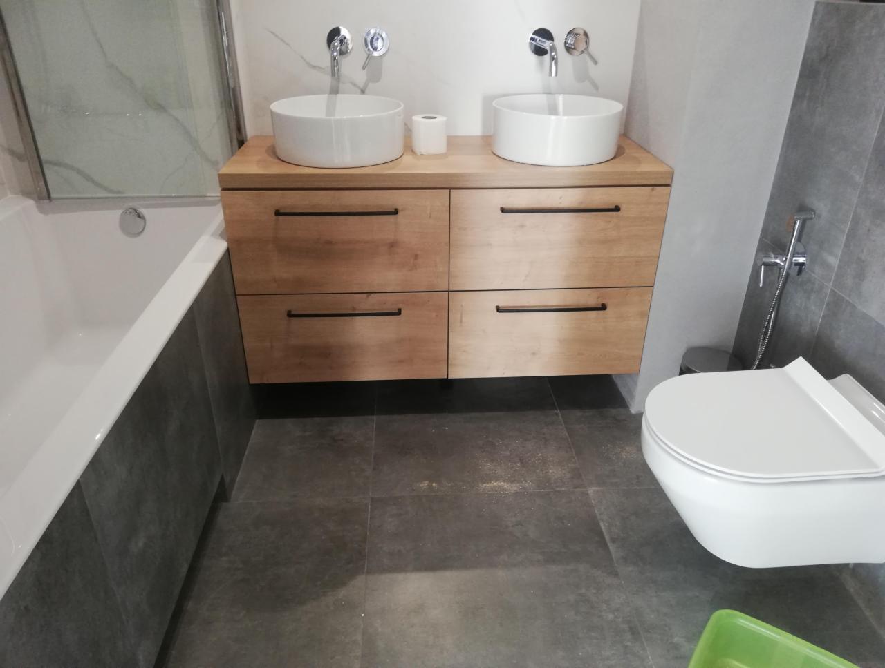 Nowoczesna łazienka z dwiema okrągłymi umywalkami nablatowymi na drewnianej szafce z czarnymi uchwytami, wisząca toaleta i szare płytki podłogowe.