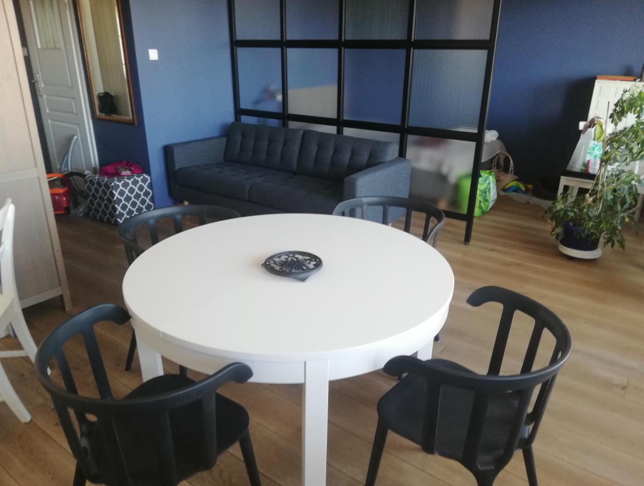 Jasne drewniane podłogi w pokoju dziennym z białym okrągłym stołem, czarnymi krzesłami, szarą sofą i niebieskimi ścianami, oddzielonymi szklaną ścianką działową.