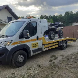 Biała laweta z żółtymi pasami i numerami telefonów przewożąca quada na platformie, zaparkowana na żwirowym podłożu przed domem i drewnianym ogrodzeniem.