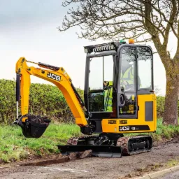 Żółta minikoparka JCB 19C-1 z łyżką pełną ziemi, operator w kamizelce odblaskowej w kabinie, na tle zielonego żywopłotu i drzewa.