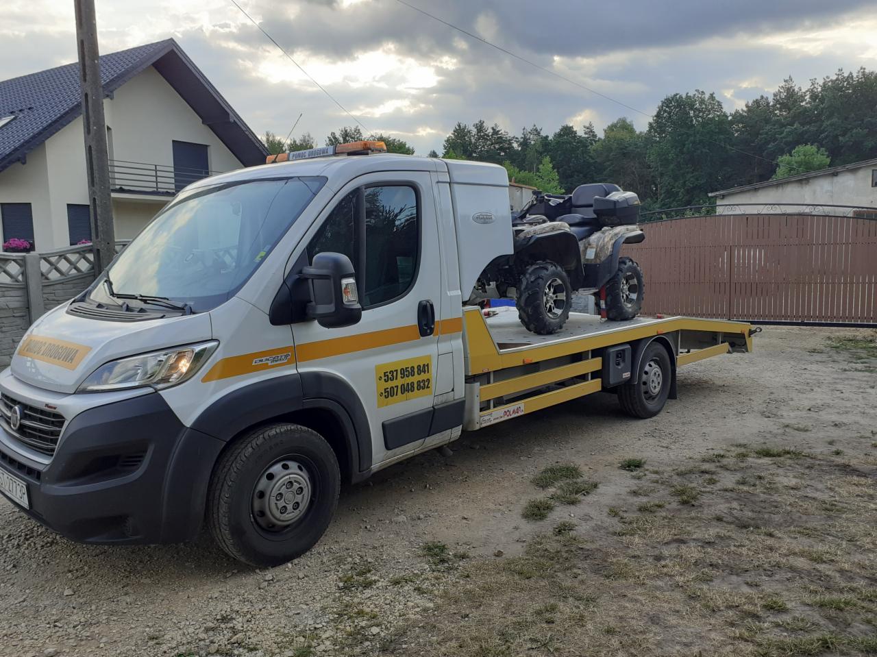 Biała laweta z żółtymi pasami i numerami telefonów przewożąca quada na platformie, zaparkowana na żwirowym podłożu przed domem i drewnianym ogrodzeniem.