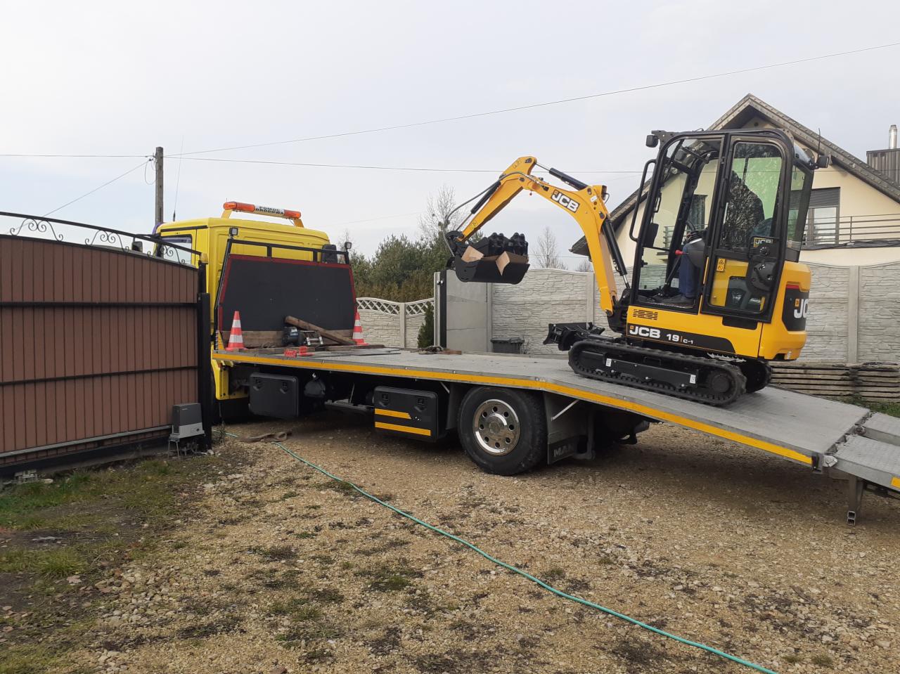 Żółta mini koparka JCB 19c-1 na platformie żółtej ciężarówki z pomarańczowym światłem ostrzegawczym, zaparkowana na żwirowym podłożu przed ogrodzeniem.