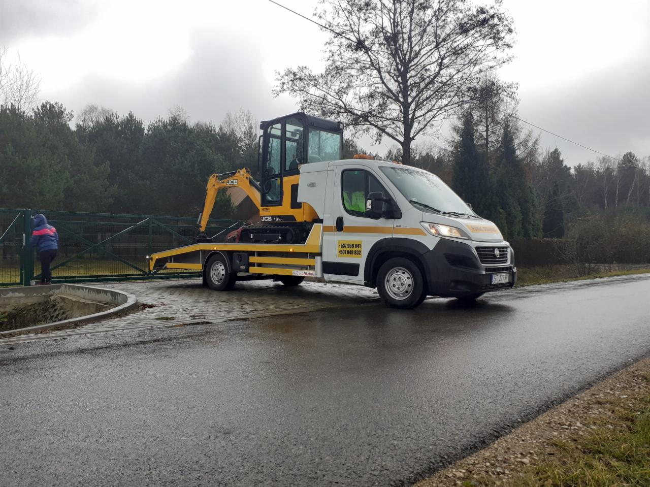 Biała laweta z żółtymi pasami przewozi małą, żółtą koparkę JCB 18z-1 na tle drzew i pochmurnego nieba. Po lewej stronie kadru osoba w niebieskiej kurtce stoi przy bramie.