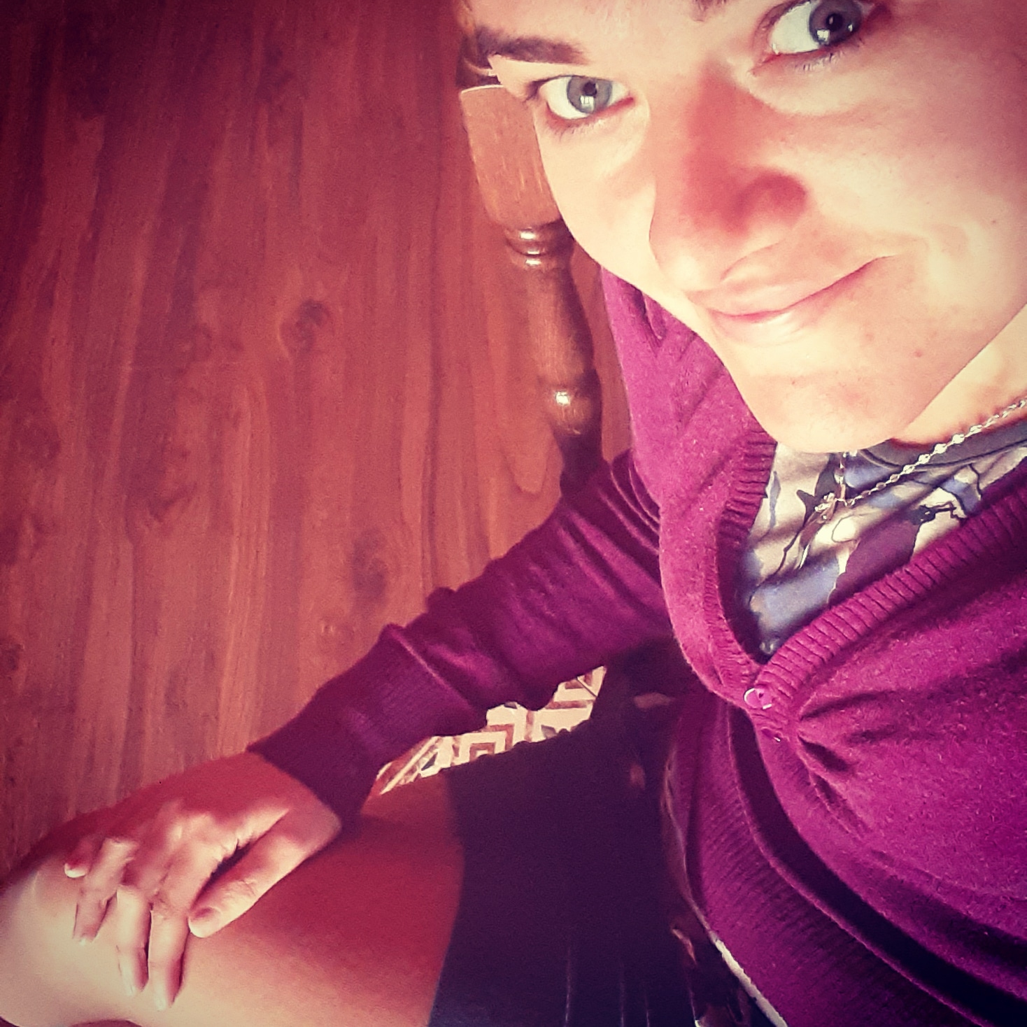 Portret kobiety z bliska, uśmiechniętej, w fioletowym swetrze, na tle drewnianej ściany. Widoczne jasne oczy i delikatny uśmiech.