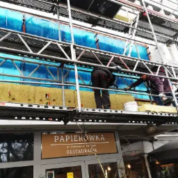 Pracownicy na rusztowaniu montują izolację termiczną na elewacji budynku, zabezpieczonej folią ochronną, nad wejściem do restauracji Papierówka.