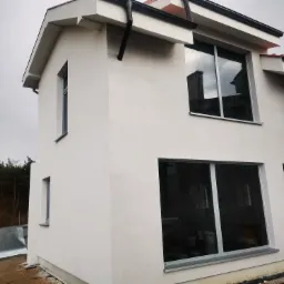 Świeżo otynkowany, dwupiętrowy dom z grafitowymi oknami i rynnami, widoczne rusztowania i blacha falista w tle.