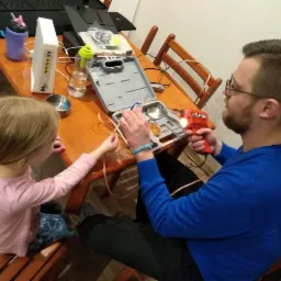 Mężczyzna z lutownicą TLP 150 łączy kabel, asystuje mu dziewczynka siedząca obok, na stole router, woda i otwarty zestaw narzędzi.