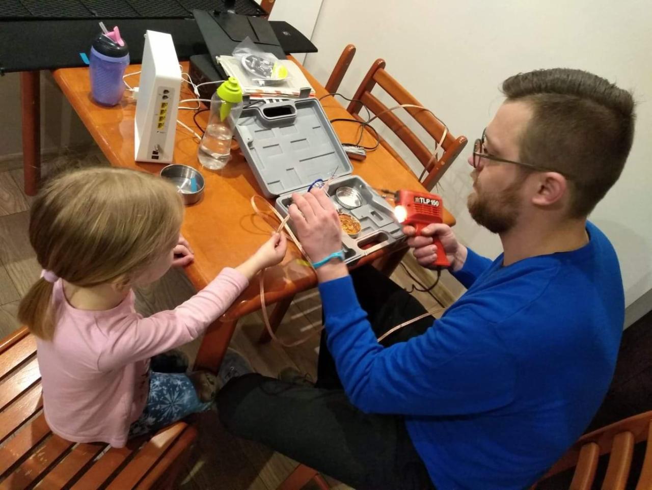 Mężczyzna z lutownicą TLP 150 łączy kabel, asystuje mu dziewczynka siedząca obok, na stole router, woda i otwarty zestaw narzędzi.