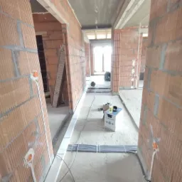 Wnętrze w trakcie budowy z widocznymi ścianami z cegły, instalacją elektryczną w puszkach i wiązkami kabli na podłodze; w tle widoczne okno i narzędzia.