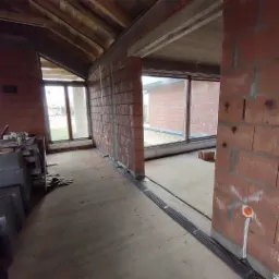 Wnętrze surowego domu w trakcie budowy: widoczne ceglane ściany z instalacją elektryczną w peszlach, duże okna i betonowa podłoga.