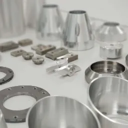 Różnorodne, precyzyjnie obrobione metalowe elementy na białym tle, w tym pierścienie z otworami, stożki i bloki, ukazujące detale frezowania i toczenia.