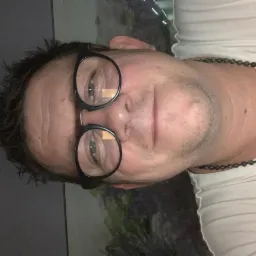 Portret mężczyzny w okularach na tle okna. Widoczne odbicie krajobrazu za szybą. Ujęcie z bliska, obrócone o 90 stopni.