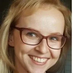 Portret uśmiechniętej blondynki w okularach o kocim kształcie, z widocznym dołeczkiem w policzku i delikatnym makijażu. Ujęcie od przodu, naturalne światło.