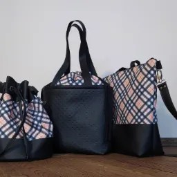 Trzy różne torby damskie: worek z sznurkiem w kratę, shopperka z czarnej perforowanej skóry i torba na ramię w kratę, wszystkie z czarnymi skórzanymi detalami, ustawione na drewnianej podłodze.