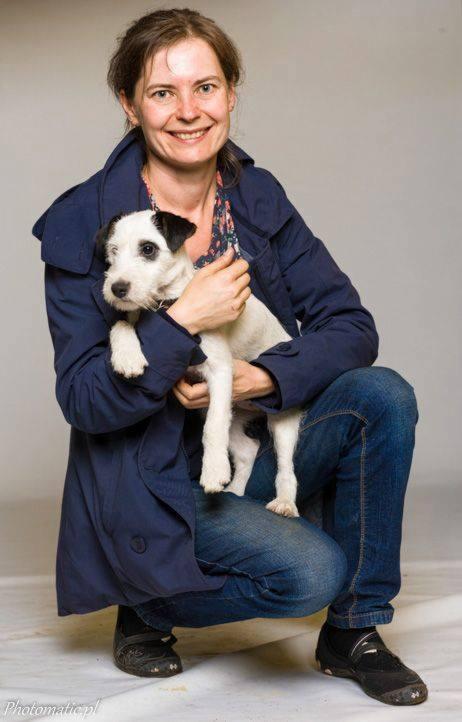 Uśmiechnięta kobieta w niebieskim płaszczu przykuca, trzymając białego szczeniaka Jack Russell Terrier z czarnymi łatami na głowie.