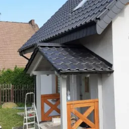 Nowoczesny dom z ciemnoszarą dachówką, widoczny fragment elewacji, drewniane balustrady ganku w stylu rustykalnym oraz drabina oparta o ścianę.