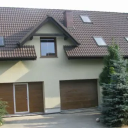 Dwukondygnacyjny dom z beżową elewacją i brązowym dachem z dachówkami, widoczne okna dachowe, garaż z bramą segmentową i drzwi wejściowe, otoczony zielenią.