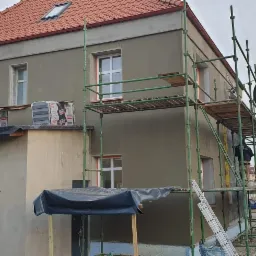 Dom w trakcie ocieplania elewacji, widoczne rusztowania, worki z materiałem izolacyjnym na dachu budynku gospodarczego, elewacja pokryta szarym tynkiem.