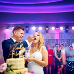 Uśmiechnięty pan młody karmi tortem otwartą buzię panny młodej podczas wesela; w tle goście i scena oświetlona fioletowymi światłami.