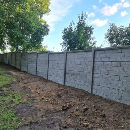 sl-service - Długi, szary mur z betonowych elementów imitujących cegłę, oddzielający trawnik od ziemi, widoczny w perspektywie z niskiego kąta.