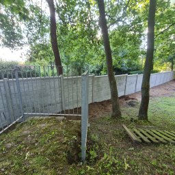 sl-service - Wznoszenie ogrodzenia: widok na szary betonowy mur i metalową siatkę w trakcie montażu, z widocznymi słupkami i wykopanym dołem pod fundament, w tle drzewa i zieleń.
