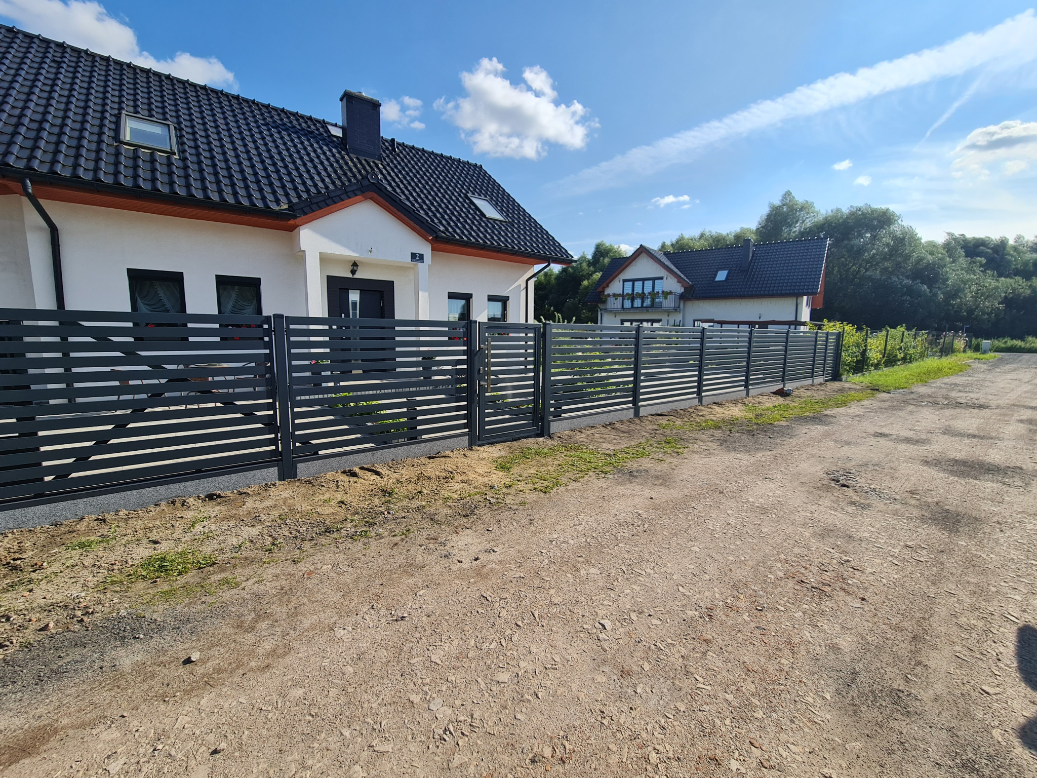 Nowoczesne poziome ogrodzenie z szarych paneli na tle domu z czarną dachówką i błękitnego nieba z chmurami.