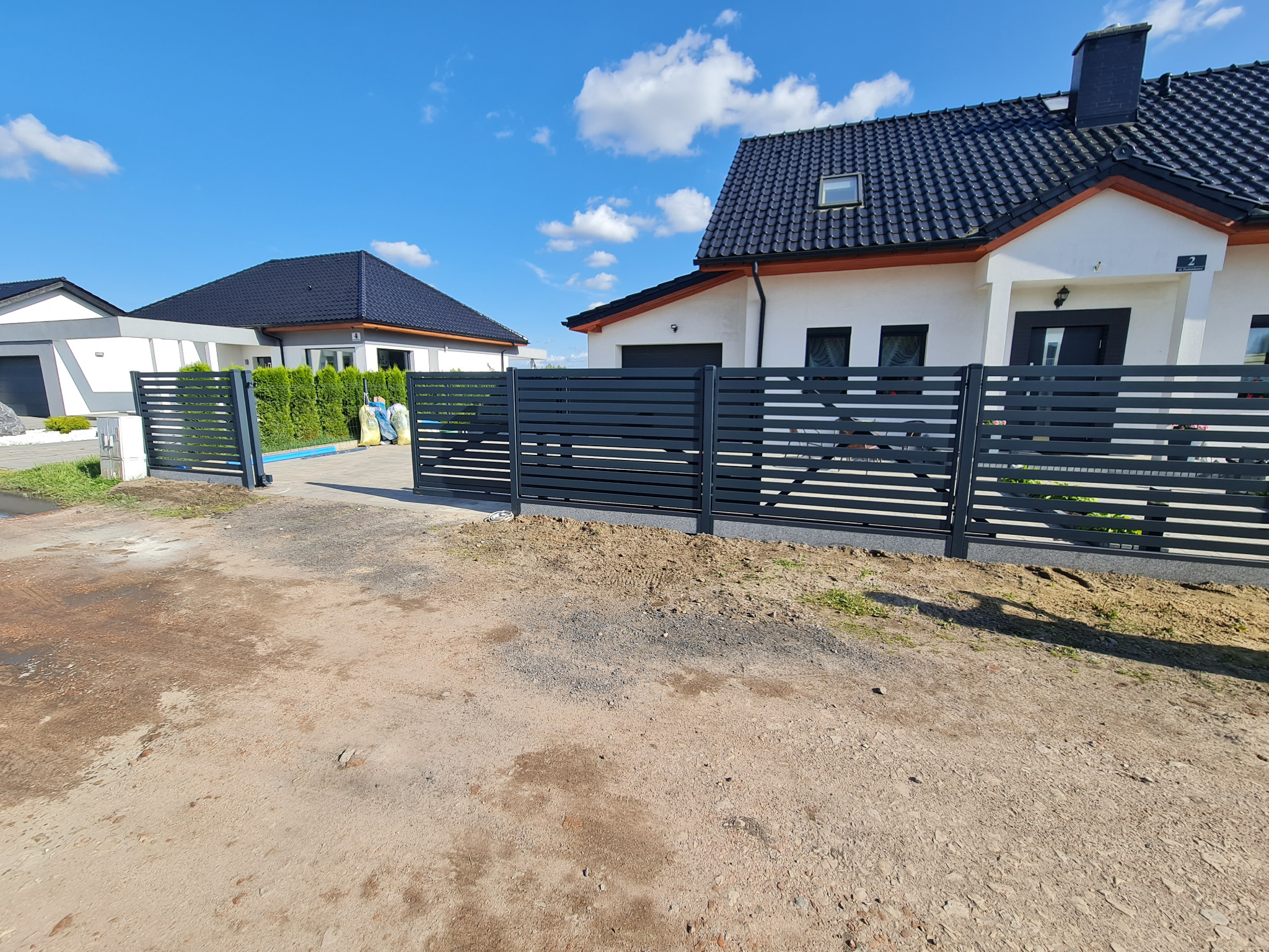 Nowoczesne, poziome przęsła ogrodzeniowe w kolorze grafitowym przed nowo wybudowanymi domami z ciemną dachówką, widok na otwartą bramę wjazdową.