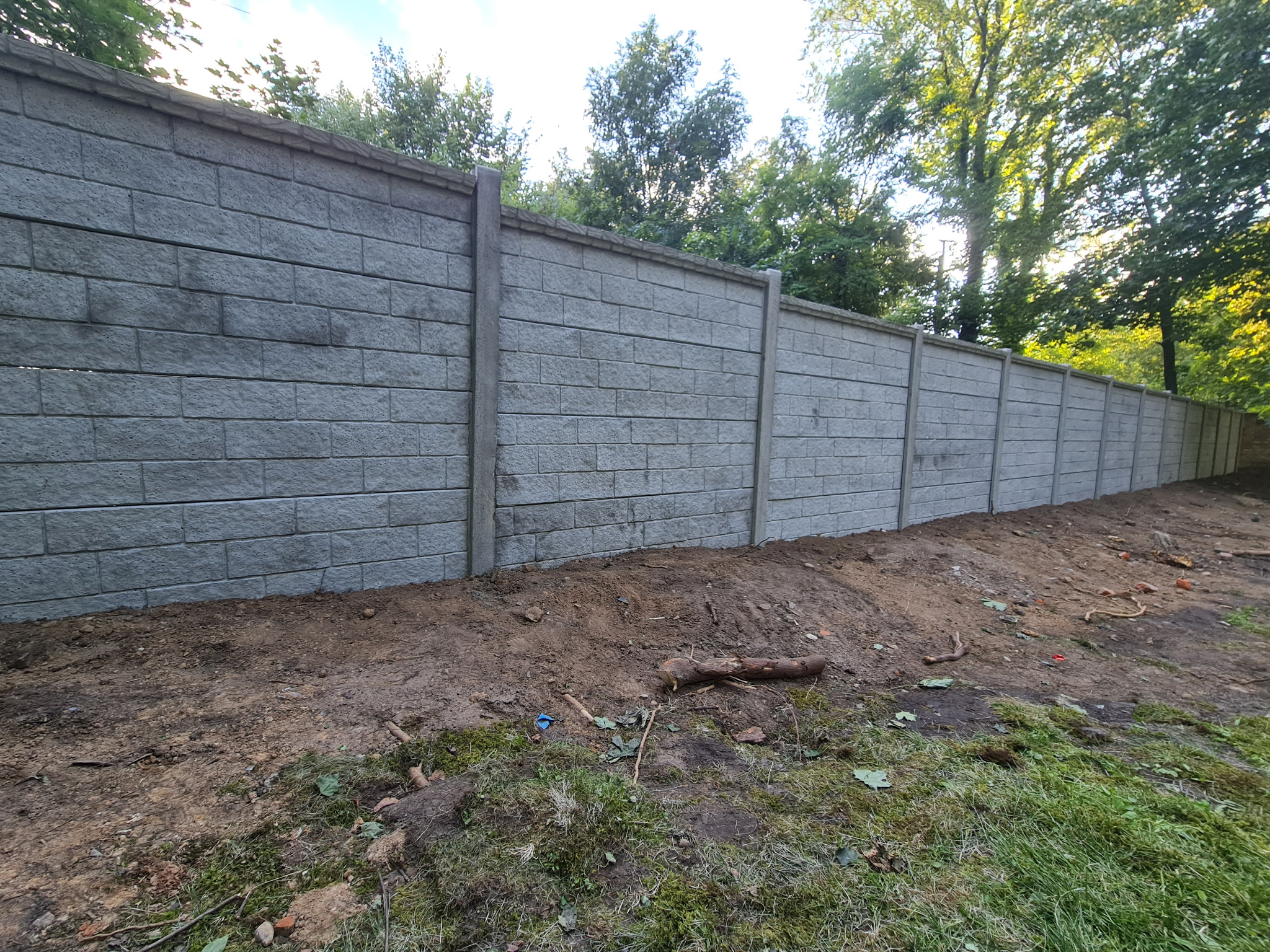 Długi, szary mur z betonowych bloczków oddzielający teren, widoczne słupki wzmacniające konstrukcję, ziemia po montażu jeszcze nie wyrównana, w tle zieleń drzew.