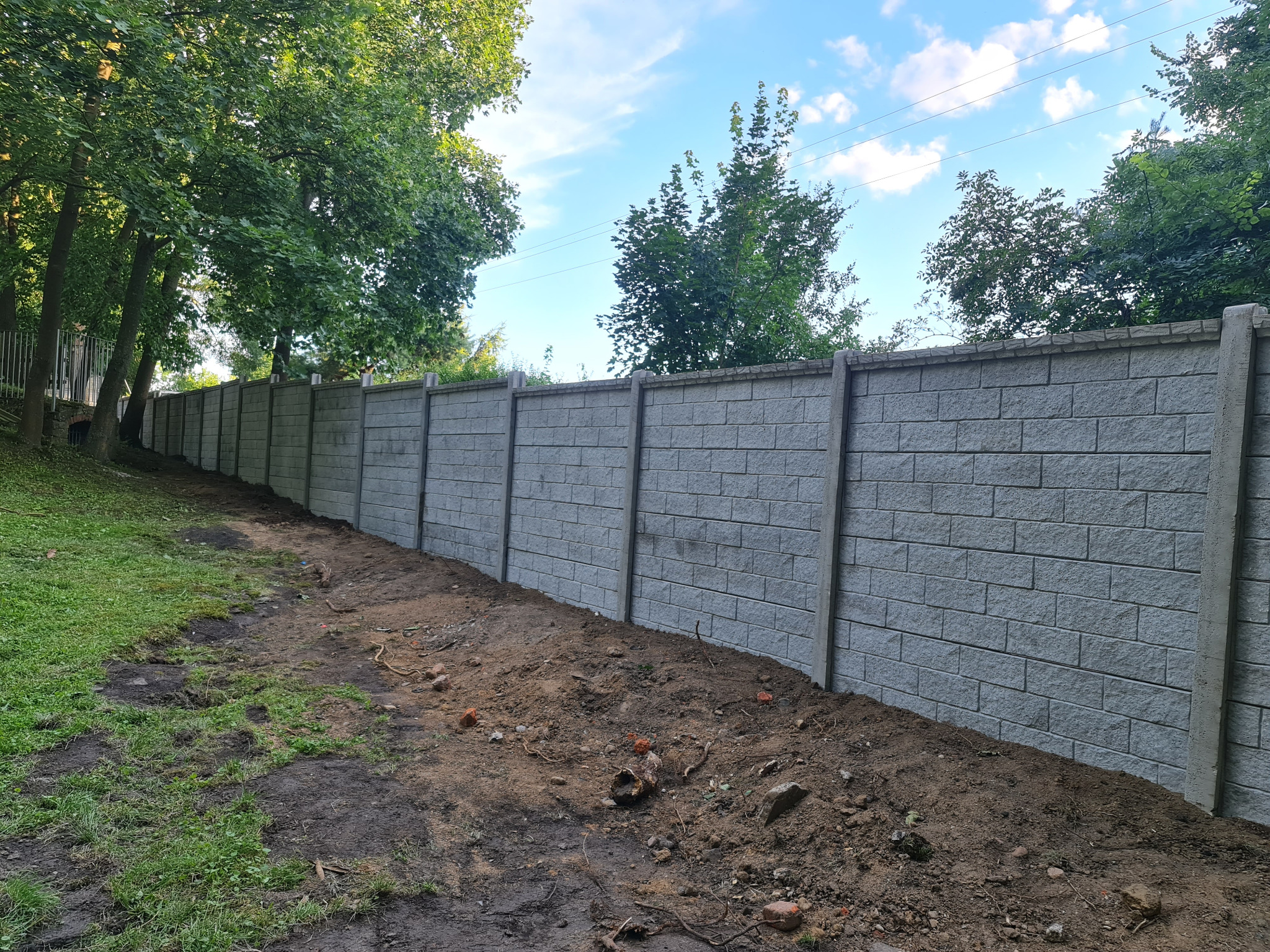 Długi, szary mur z betonowych elementów imitujących cegłę, oddzielający trawnik od ziemi, widoczny w perspektywie z niskiego kąta.