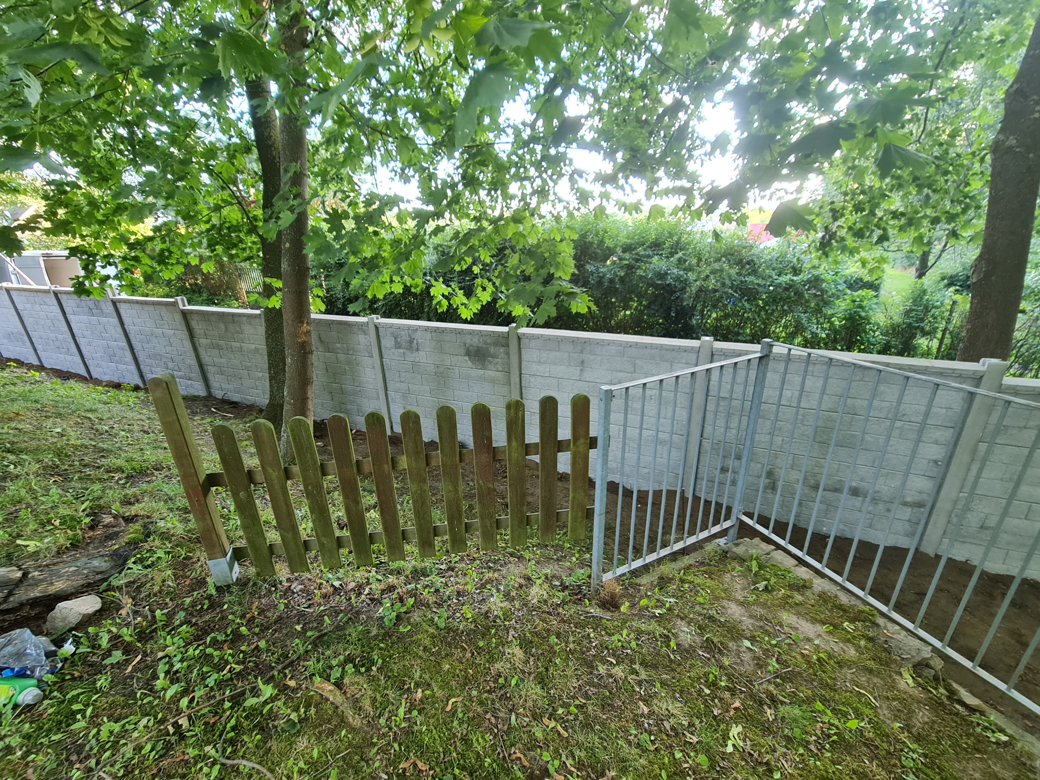 Fragment posesji z nowym, szarym ogrodzeniem z prefabrykatów betonowych i dwoma różnymi typami furtek: starą, drewnianą i nową, metalową, z widocznymi śladami prac ziemnych.