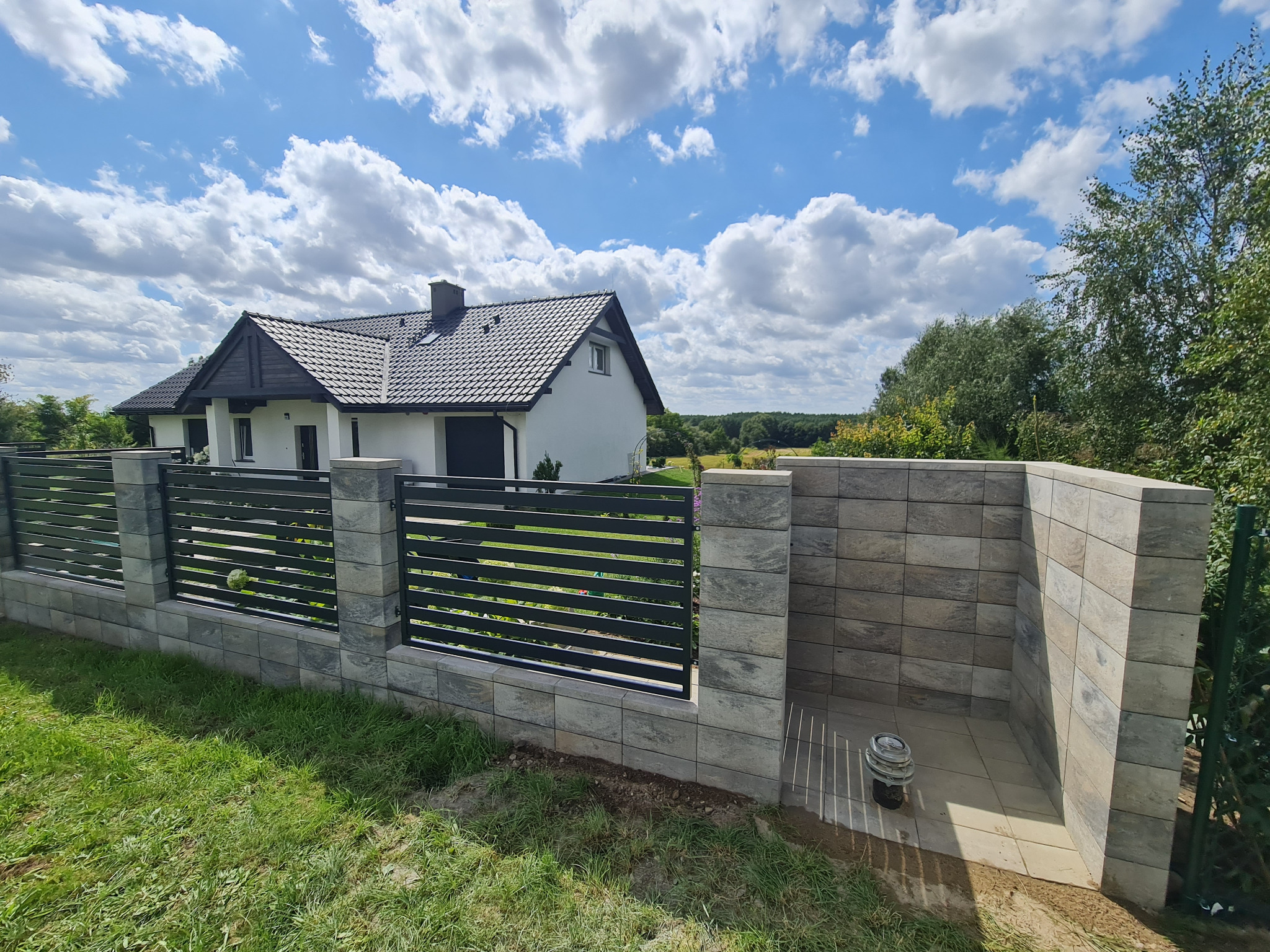 Nowoczesne ogrodzenie z poziomych paneli stalowych i betonowych słupków, z domem w tle pod błękitnym niebem z chmurami.