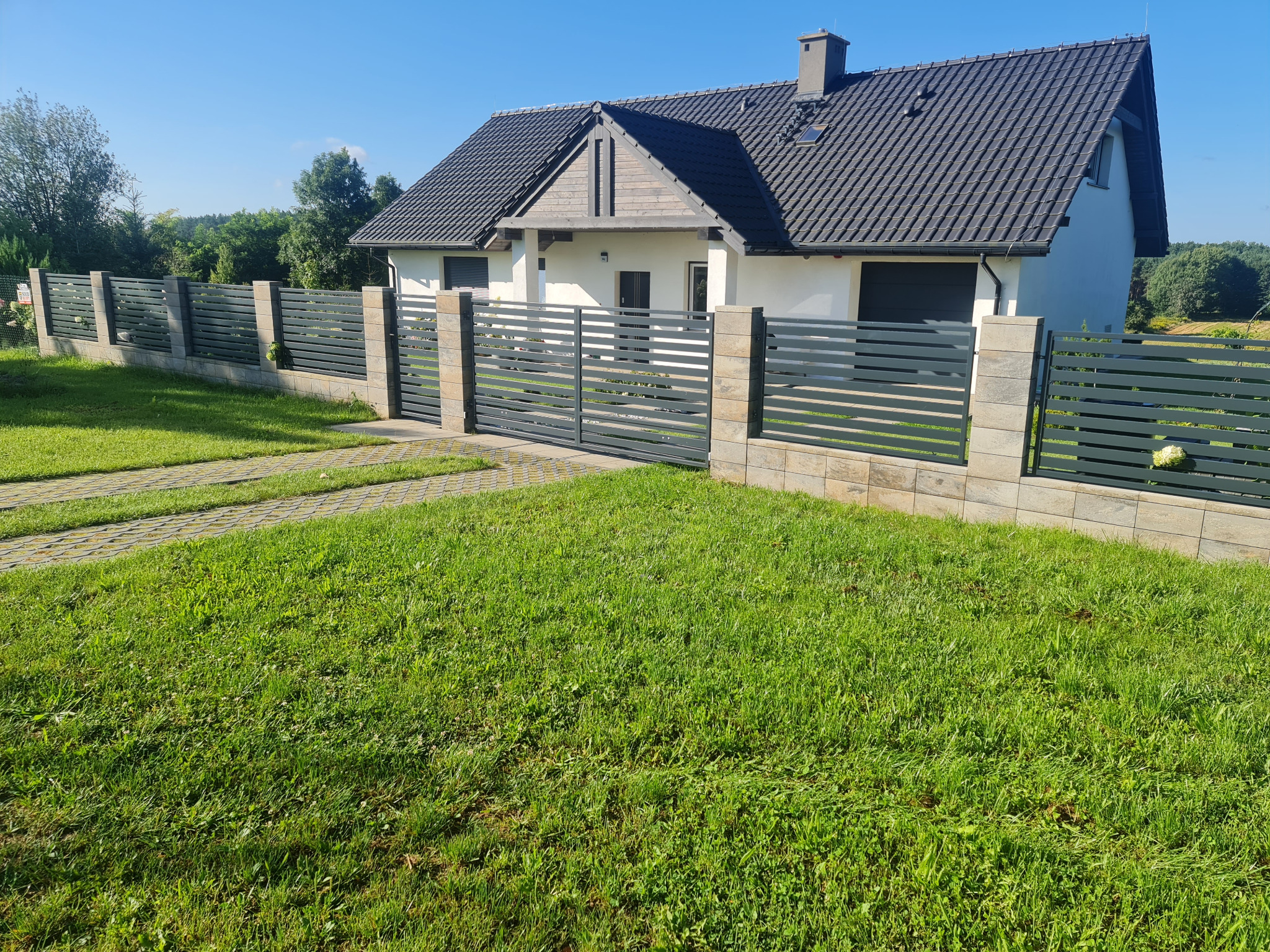 Nowoczesna posesja z szarym, poziomym ogrodzeniem z przęsłami i słupkami z kamienia dekoracyjnego, brama wjazdowa w kolorze ogrodzenia, trawnik i dom jednorodzinny w tle.